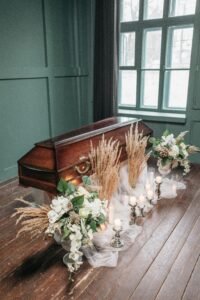 A serene indoor funeral setting featuring a wooden coffin, candles, and delicate floral decorations.