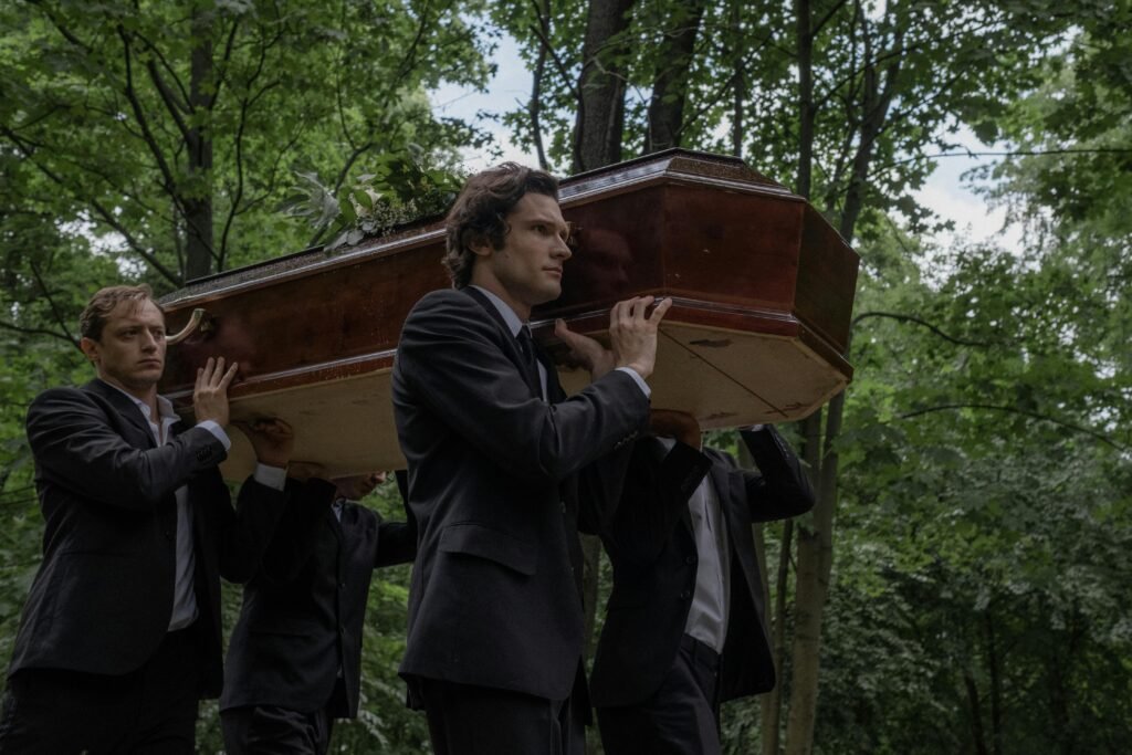 Men in black suits solemnly carry a coffin through an outdoor forest setting.